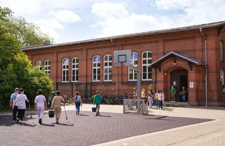 zeigt den Schulhof der Lukas-Backmeister-Schule mit Kindern, die Müll aufsammeln, im Hintergrund das rote Backsteingebäude.