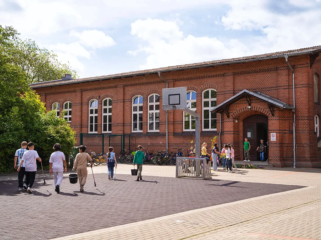 zeigt den Schulhof der Lukas-Backmeister-Schule mit Kindern, die Müll aufsammeln, im Hintergrund das rote Backsteingebäude.