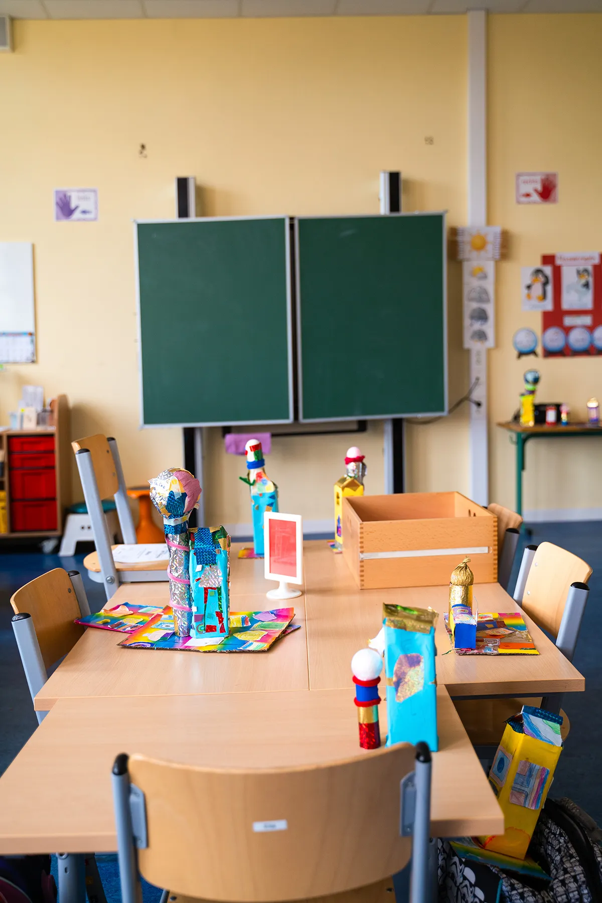 zeigt einen Klassenraum. Im Hintergrund eine Tafel, im Vordergrund ein Tisch mit Bastelarbeiten.