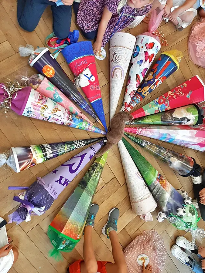 zeigt Schultüten auf dem Boden liegend, kreisförmig, angeordnet. Drumherum sitzen Kinder.