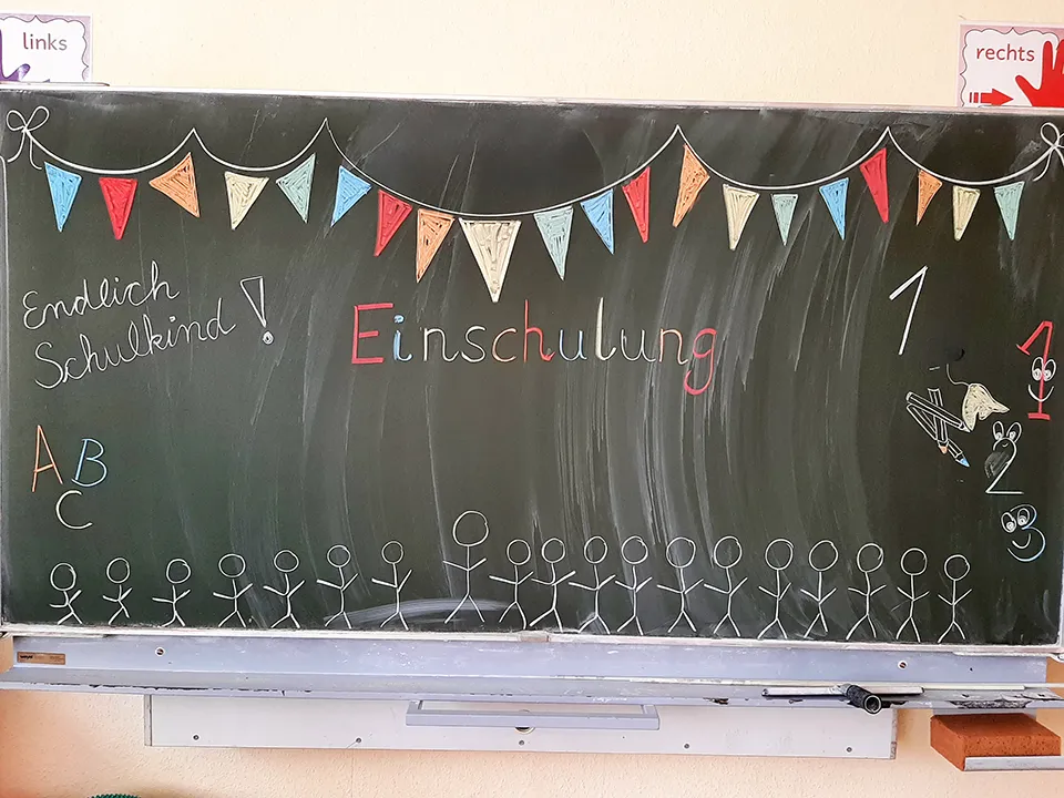 zeigt eine Schultafel mit dem Wort Einschulung. Darüber hängen bunte Fähnchen.